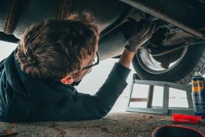 Homem inspeciona a parte inferior de um carro elevado na oficina, verificando possíveis ajustes de alinhamento e balanceamento.